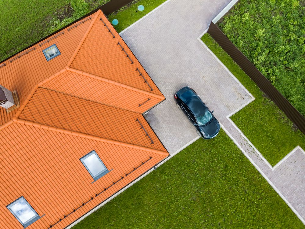 Aerial view of a house with a metal shingle roof, showcasing orange tiles, two windows, and a parked black car in the driveway, surrounded by green grass and a fence.
