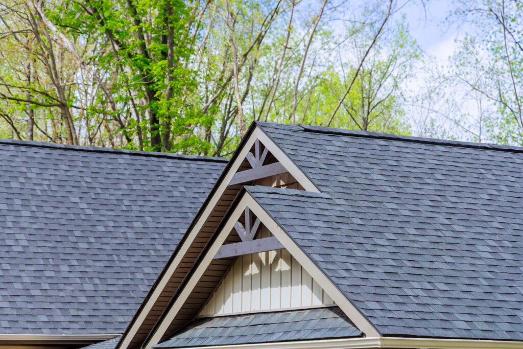 Asphalt shingle roofing on a newly constructed home, featuring a steep pitch, detailed gable design, and surrounded by lush green trees.