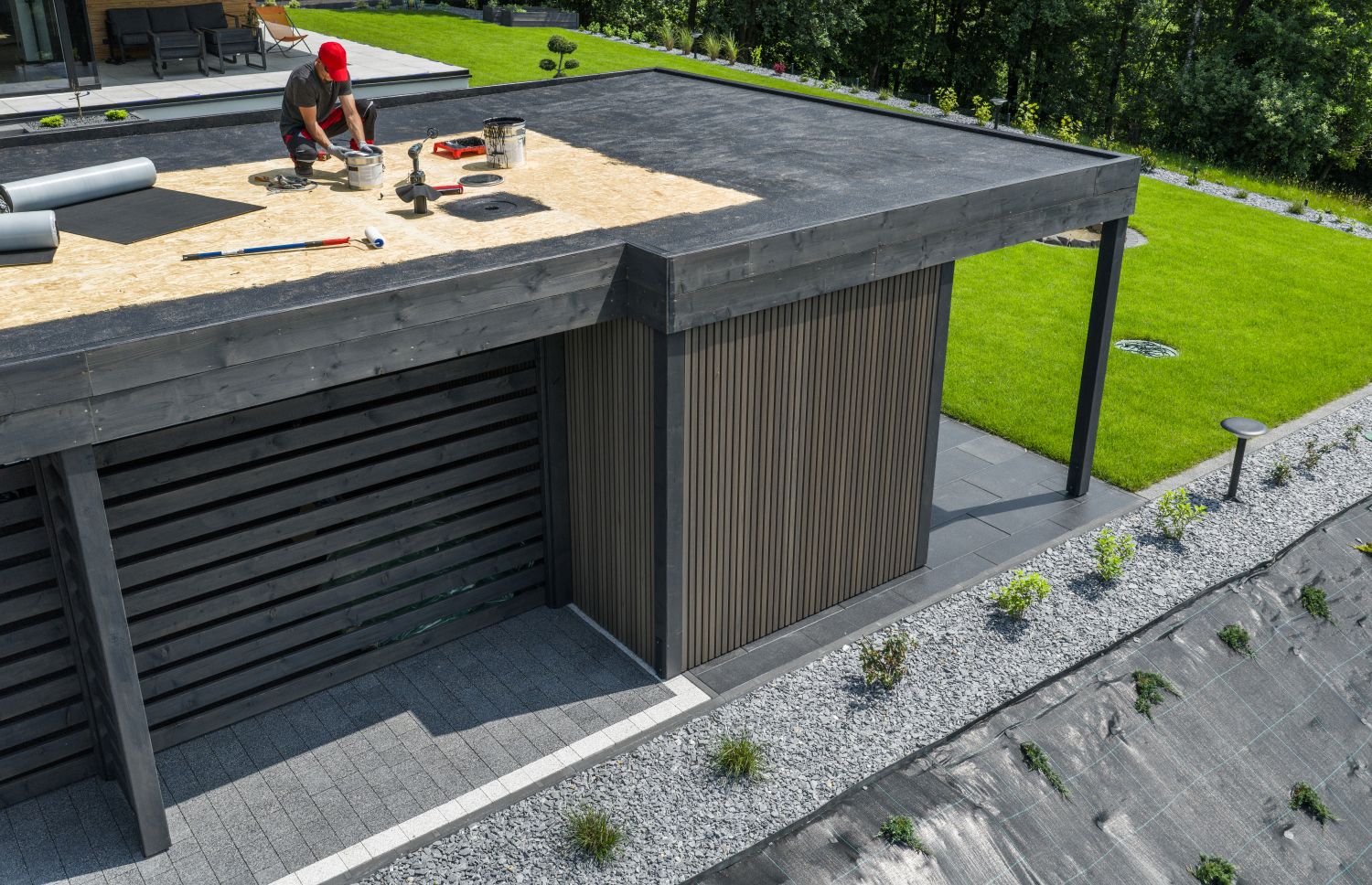 Workers performing roof maintenance on a modern flat roof, utilizing tools and materials for installation, surrounded by a landscaped area.